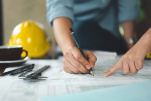 Close-up of an engineer’s hands using a pencil to review detailed plumbing blueprints and technical drawings on a desk.