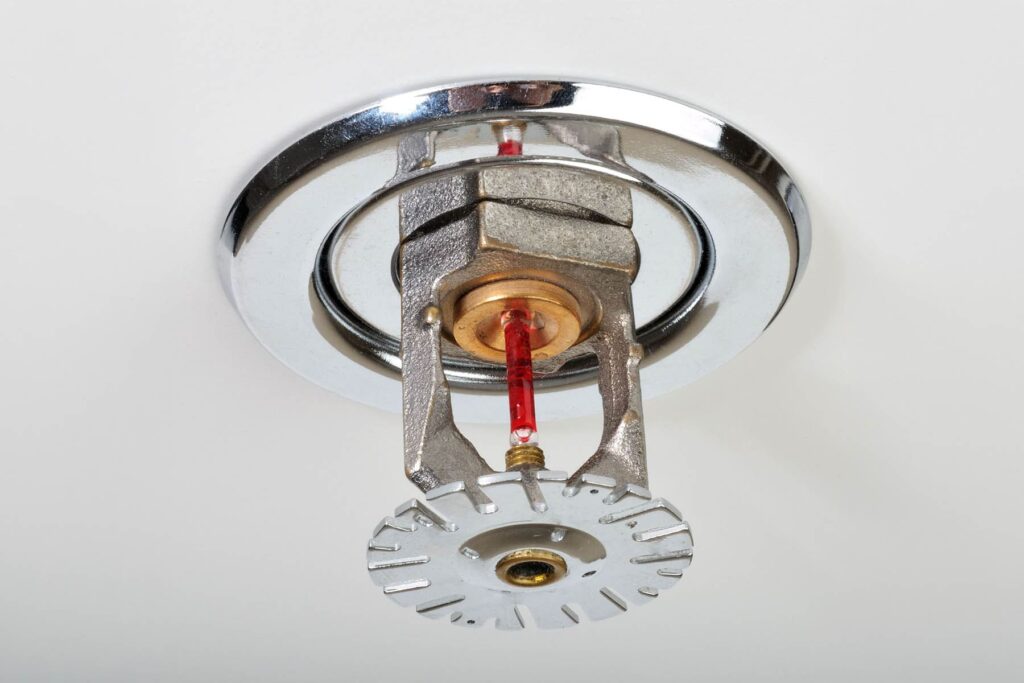 A detailed close-up of a chrome-finished fire sprinkler head with a red heat-sensitive glass bulb, installed in a white ceiling.