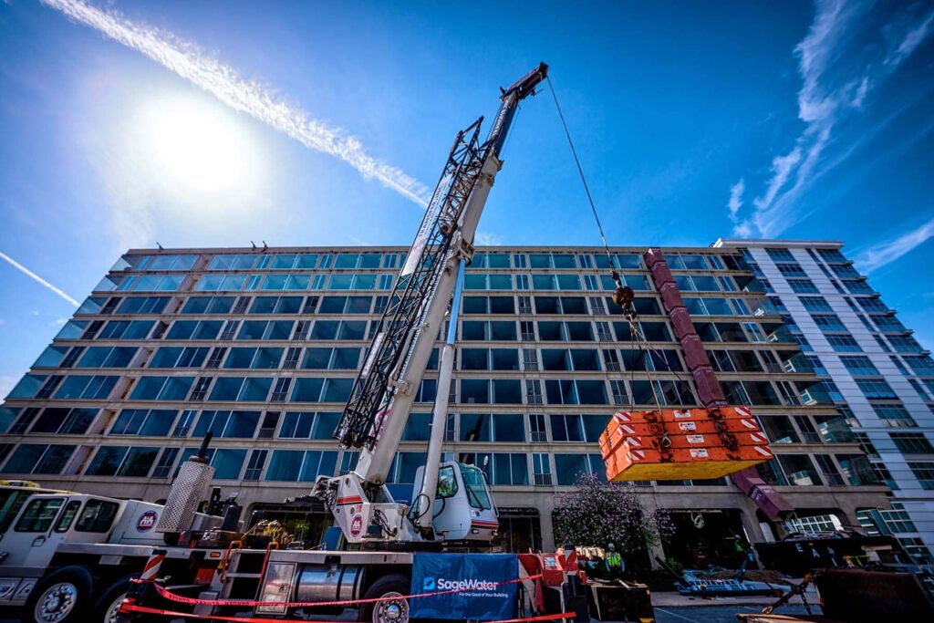 A large crane lifts a heavy equipment component at a multi-story building site with a SageWater banner in the foreground.