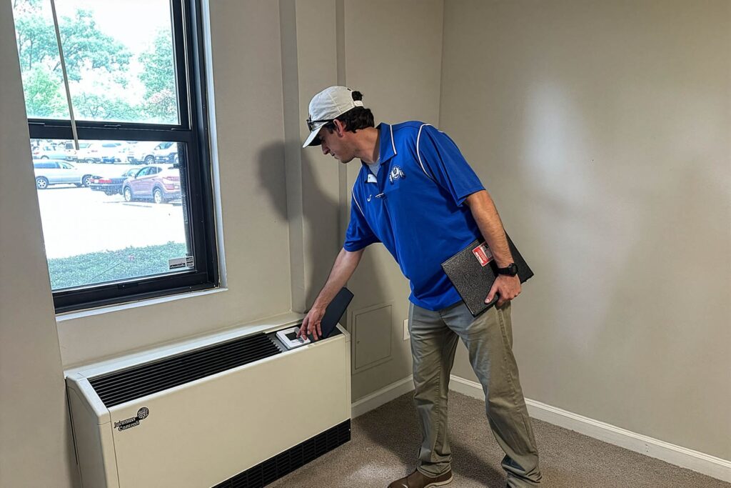 A technician in a blue SageWater shirt inspects a Johnson Controls PTAC heating and cooling unit inside a residential apartment.
