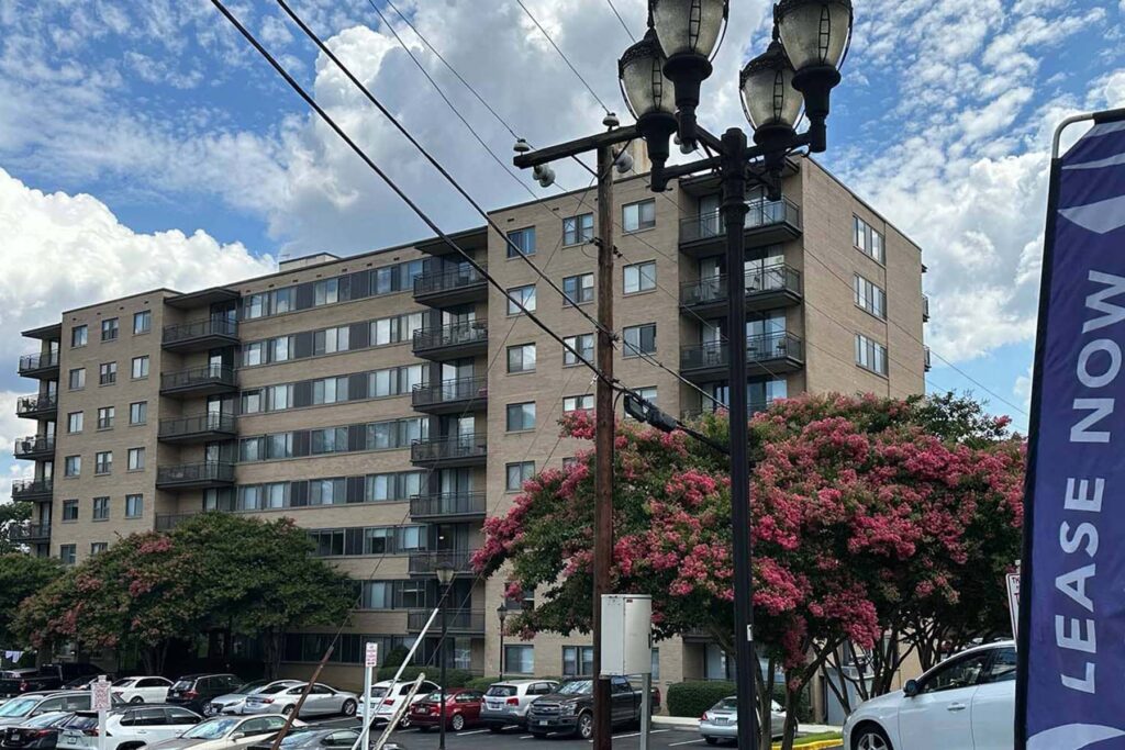 Exterior view of a high-rise multifamily apartment building with a "Lease Now" banner and flowering trees in the foreground.