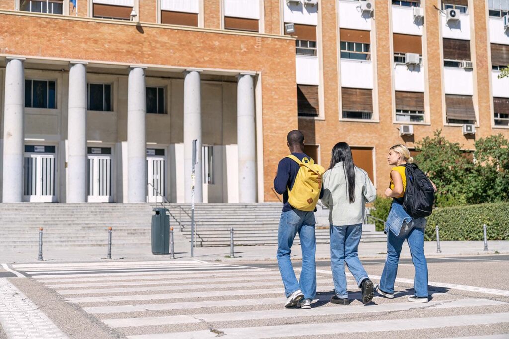 Three students with backpacks walking toward a large brick university dormitory or student housing complex.