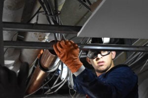 A construction worker wearing orange protective gloves and safety glasses installs electrical wiring and conduit within a ceiling's metal framing.